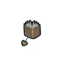 Macon & Lesquoy open book lapel pin hand-embroidered in silver and gold thread with a hanging heart charm, on a plain white background